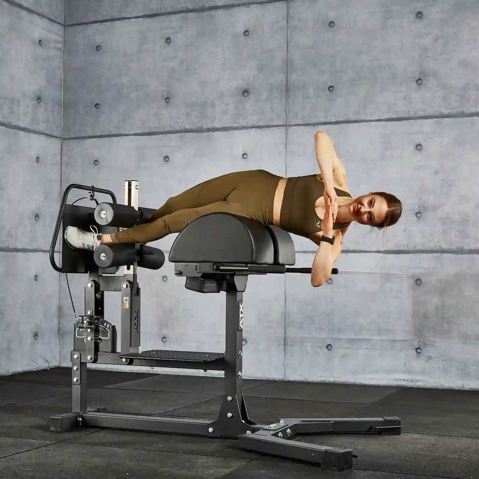 A person in workout attire is performing a side plank on a hyperextension bench, next to a glute machine, in a gym. The background features gray concrete walls and black flooring.
