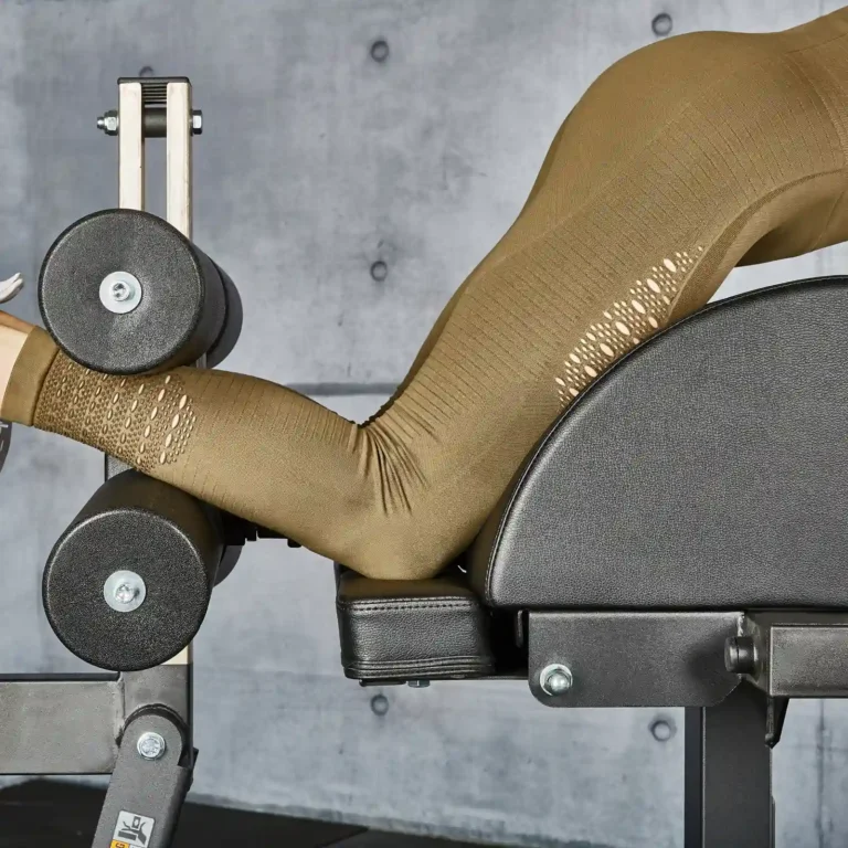 A person wearing brown leggings is using a GHD gym machine for back extensions. The focus is on their lower body and the equipment against a gray concrete wall.