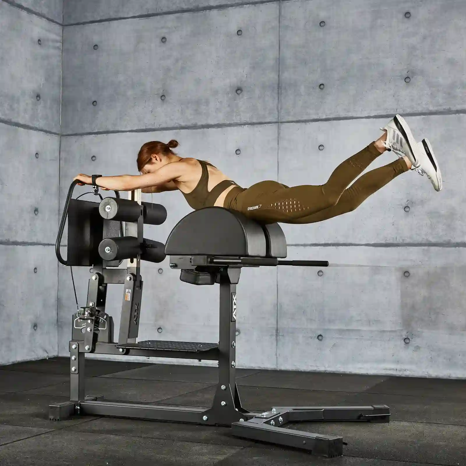 A person in athletic clothing performs a glute extension on a hyperextension bench in a gym. The background features a concrete wall with visible seams.