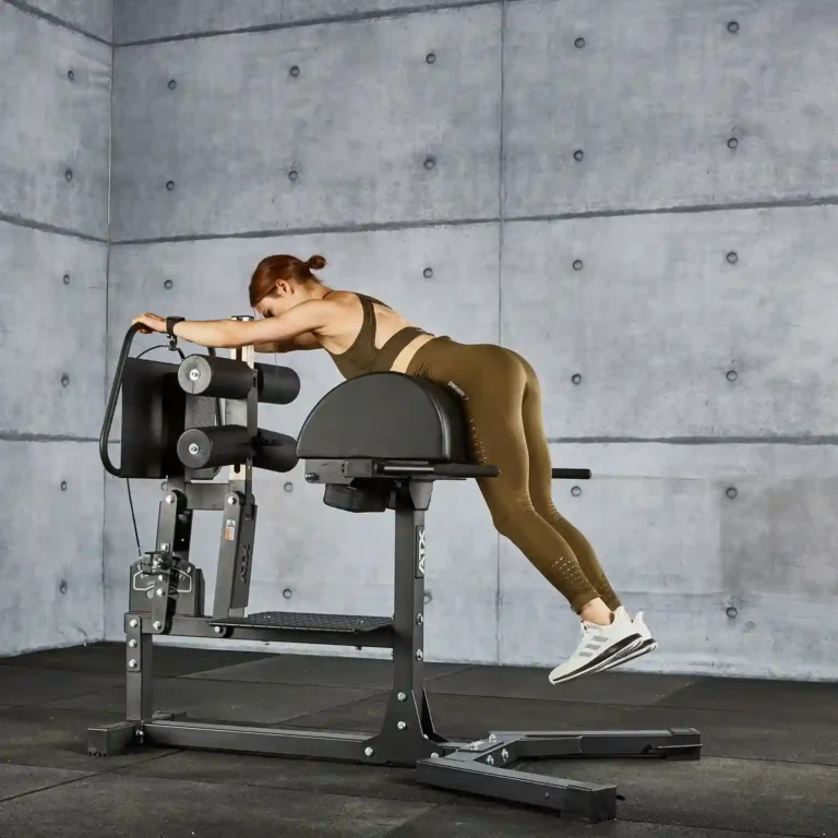 A woman in a brown workout outfit uses a GHD gym machine for exercise in a gym with concrete walls. She is positioned face-down and stretching forward, wearing white sneakers.
