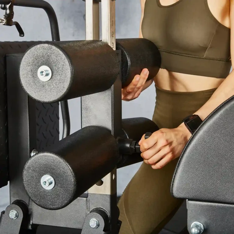 A person in a green sports outfit adjusts the settings on a gym leg press machine, ensuring their technique complements their routine on the glute and ham raise machine. Their watch is visible on their wrist, and cylindrical weights are part of the machine in the foreground.