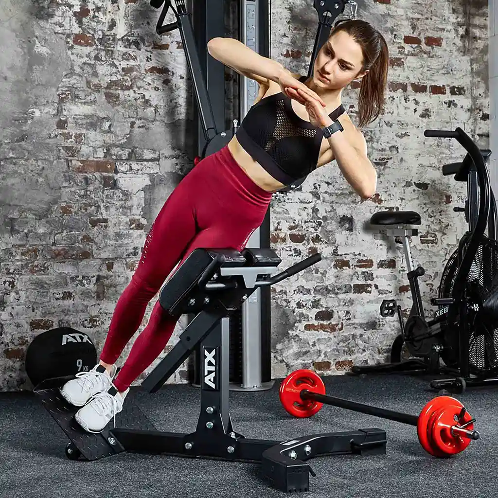 A person, dressed in a black sports top and red leggings, exercises on an ATX® Hyperextension Machine 650 in a gym. A red and black barbell lies on the floor nearby. The gym, featuring exposed brick walls, is equipped with various fitness equipment.