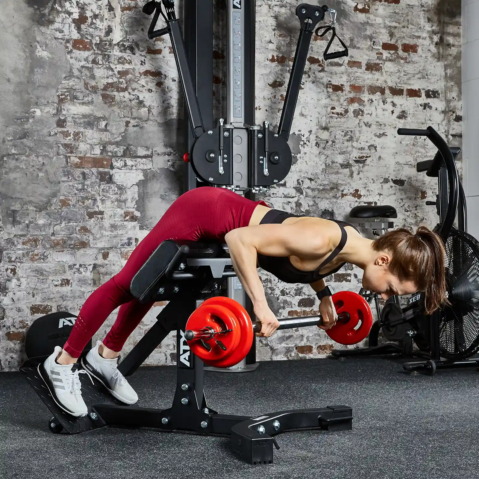 A person in a red sports outfit is performing a barbell bent-over row on a gym bench. Behind them, the gym, with its exposed brick walls and various fitness equipment, features an ATX® Hyperextension Machine 650, adding to the dynamic workout setting.