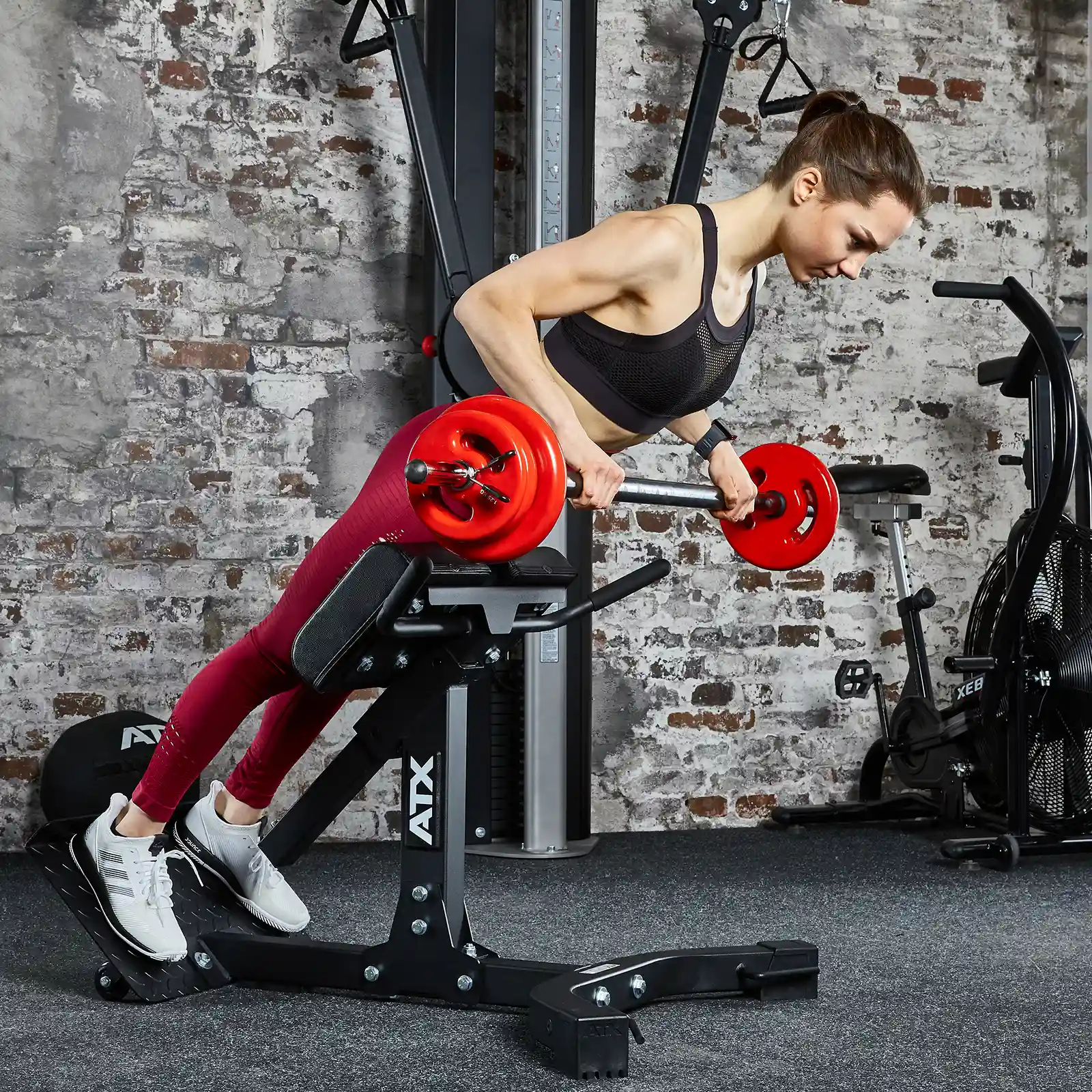 In a fitness studio, an individual performs a barbell row exercise on an incline bench, wearing a black sports top and red leggings. In the background, the ATX® Hyperextension Machine 650 is positioned against the brick wall, accompanied by various pieces of gym equipment.