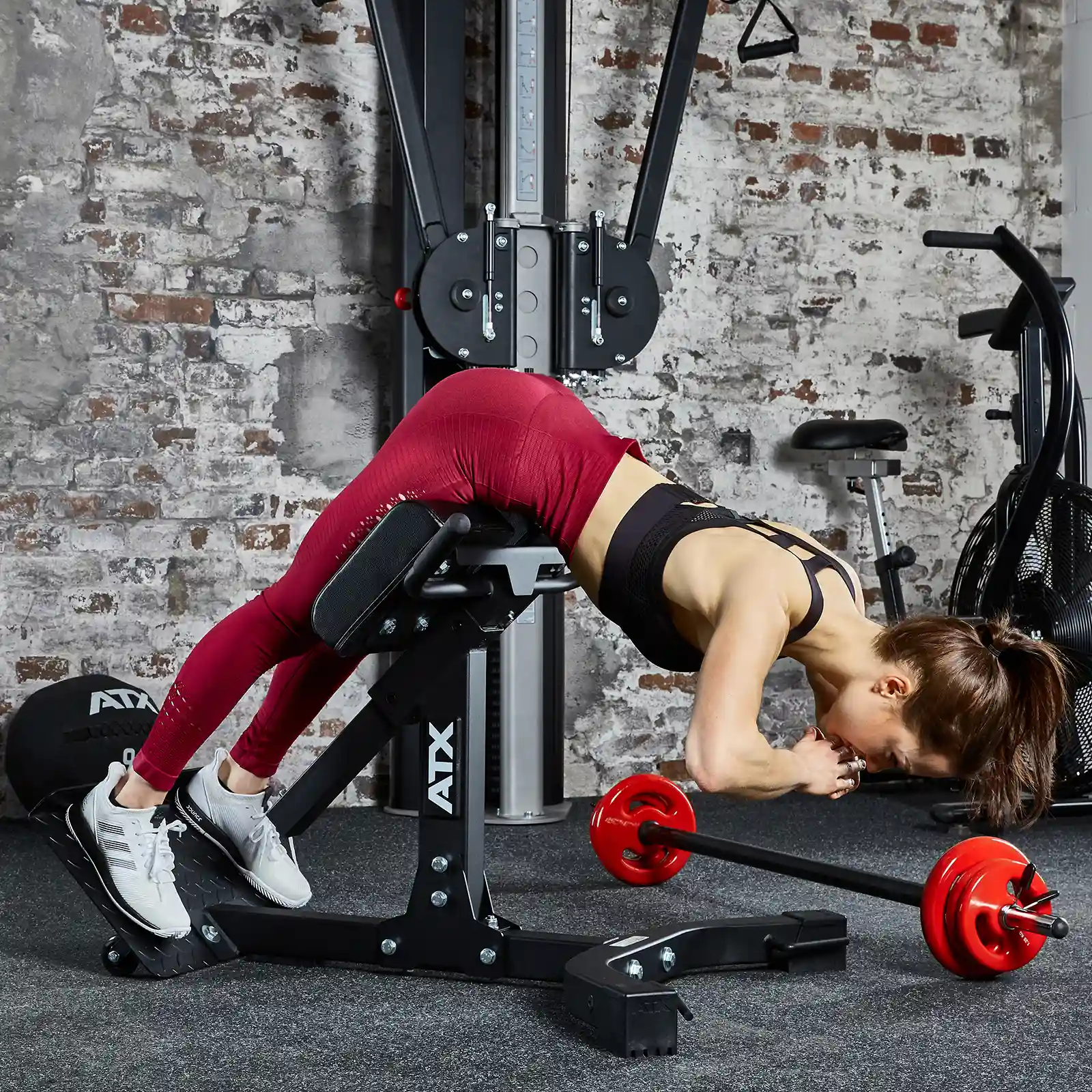 A woman in athletic attire is working out on the ATX® Hyperextension Machine 650 at the gym. She's wearing a black top, red leggings, and white sneakers. Close by, there's other gym equipment and a barbell with red weights placed against a brick-patterned wall.