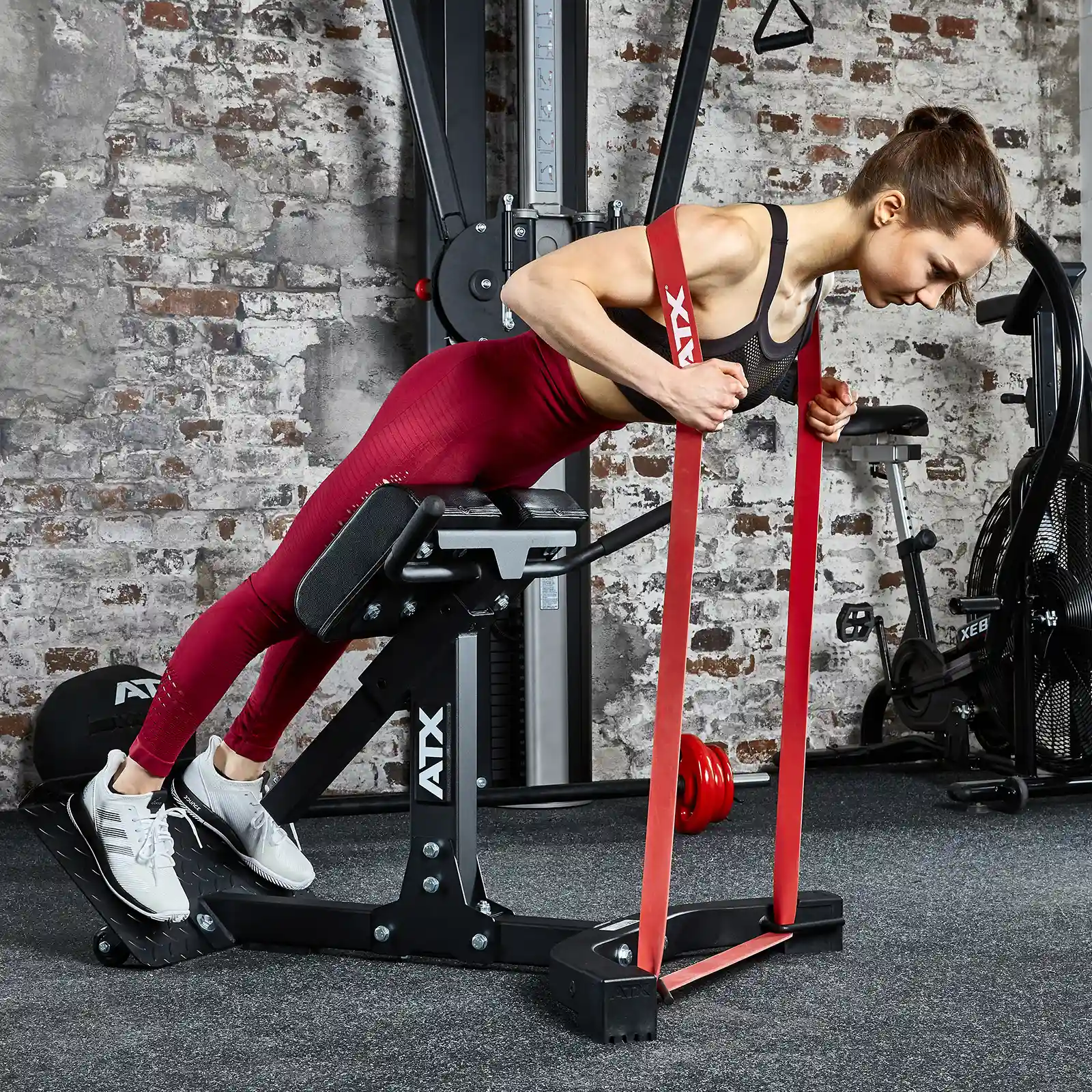 Using the ATX® Hyperextension Machine 650 in the gym, a person performs a back exercise with a resistance band. They are dressed in red leggings and a black sports top, set against a backdrop of brick walls and surrounding workout equipment.