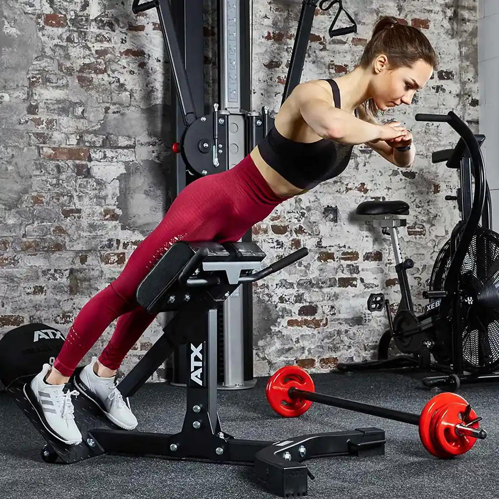 A woman in a gym performs back extensions on the ATX® Hyperextension Machine 650. She is wearing a black sports bra, red leggings, and white sneakers. A red barbell rests on the floor nearby against a backdrop of a brick wall and gym equipment.