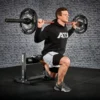 Wearing a hoodie and shorts, a person performs a barbell lunge on the ATX® Split Squat Stand, set on a black padded surface in front of a brick wall, with the barbell and equipment showcasing the ATX logo.