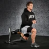 In a gym setting with a gray brick wall backdrop, someone in a black hoodie and shorts uses an ATX® Split Squat Stand to perform split squats, emphasizing balance and strength with one foot on the stand.