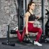 A woman in athletic gear uses the ATX® Split Squat Stand at a gym, performing a kneeling split squat while holding a kettlebell. She wears a black sports bra and red leggings, with gym equipment and a brick wall in the background.