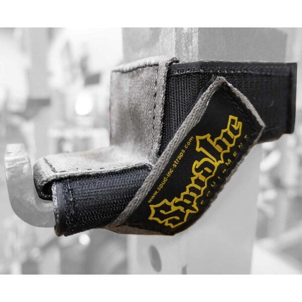 Close-up of a Spud Inc 5R Barbell Shield, black and gray with Spud Inc. in yellow, looped on a metal rack with a hook. Blurred gym equipment in the background.
