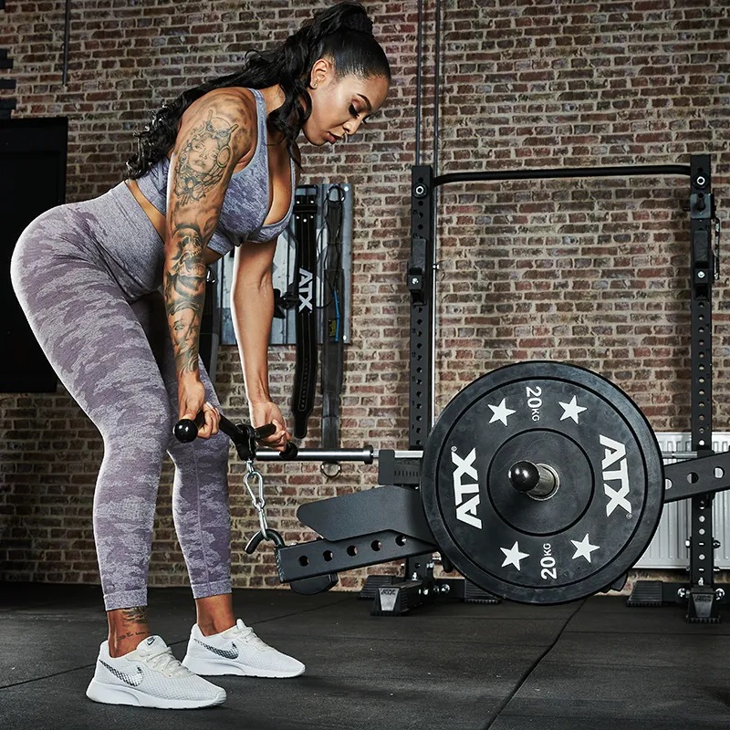 A woman in athletic wear uses the ATX® Belt Squat Option in a gym with brick walls. She is focused, gripping the equipment with both hands, and 20 kg weight plates are visible.