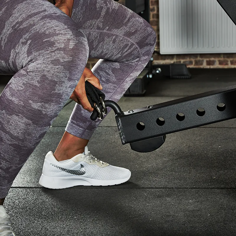 A person in gray patterned leggings and white Nike sneakers attaches a cable handle to the ATX® Belt Squat Option, preparing for a workout on a rubber gym floor.