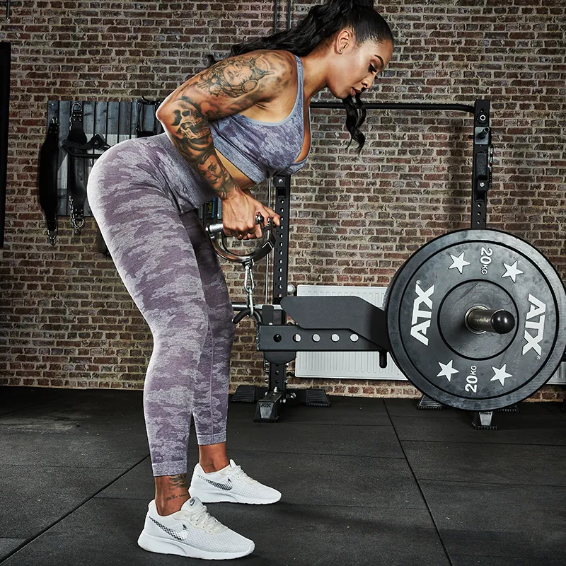 A woman in a purple workout outfit uses the ATX® Belt Squat Option in the gym, gripping the handle with both hands. Weight plates and gym equipment are visible in the background.