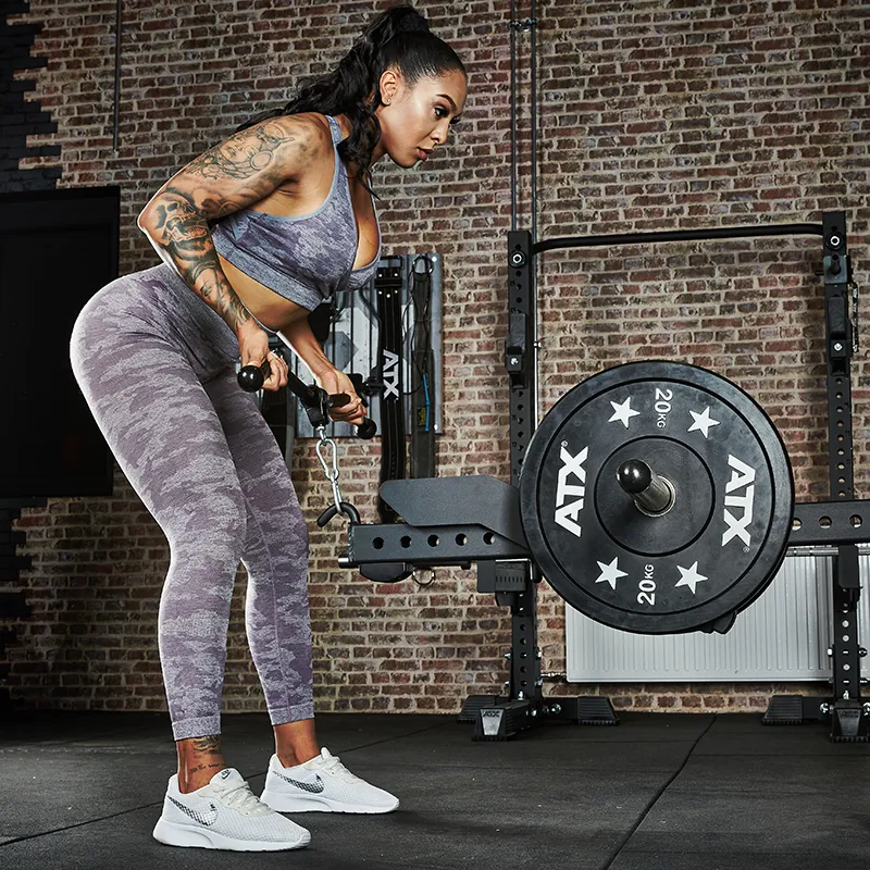 A woman wearing athletic wear uses the ATX® Belt Squat Option in a gym with brick walls, lifting weights with focus. Tattoos decorate her arms as she works out intensely.