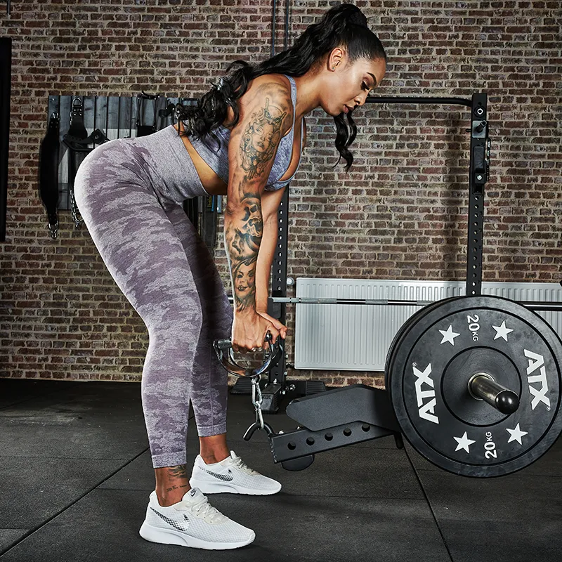 A woman uses the ATX® Belt Squat Option in a gym with brick walls and weightlifting equipment, wearing athletic gear as she performs her workout.
