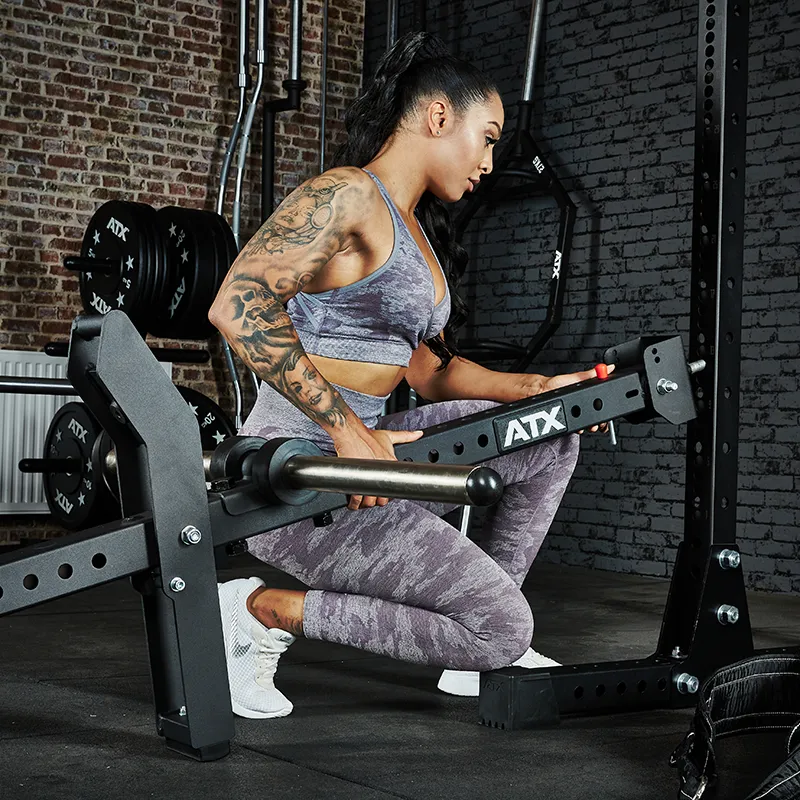 A woman in athletic wear with a ponytail and arm tattoos adjusts the ATX® Belt Squat Option in a weightlifting area featuring black weights and exposed brick walls.