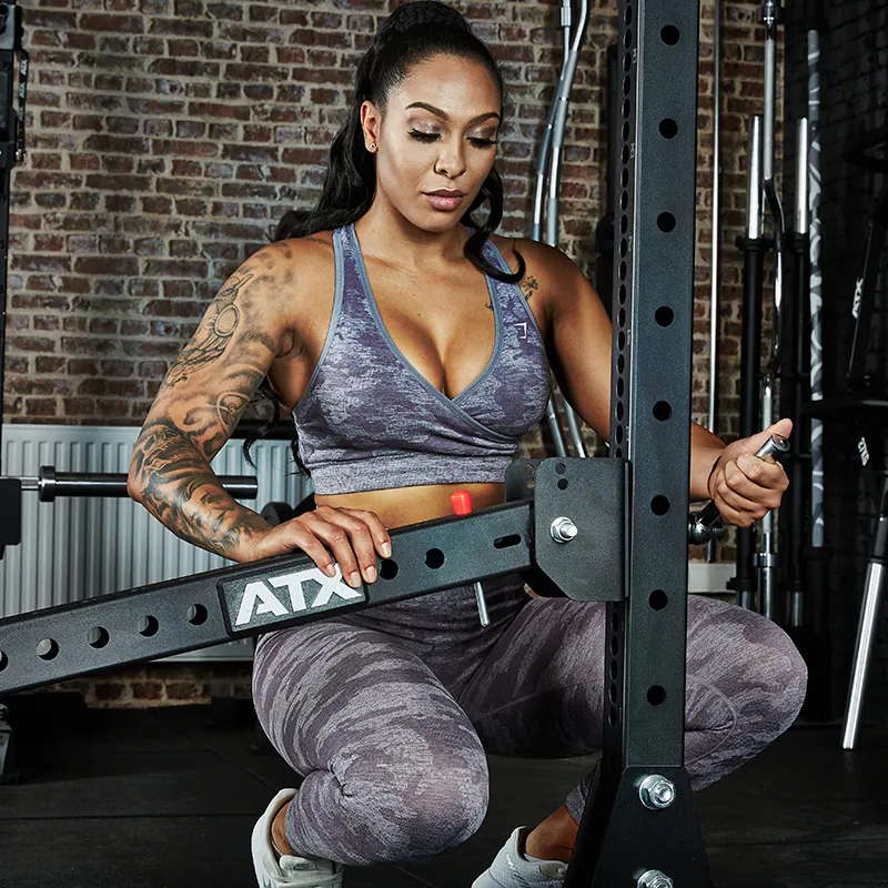 A tattooed woman in athletic wear adjusts the ATX® Belt Squat Option, focusing on securing it in a gym with other equipment and a brick wall backdrop.