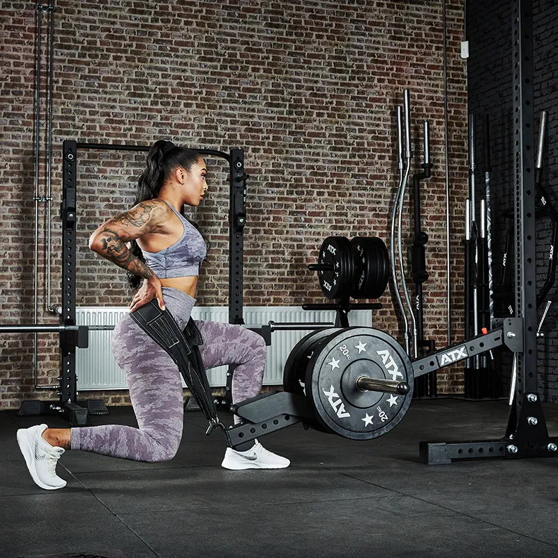 A woman in athletic wear uses the ATX® Belt Squat Option for a single-leg squat in a gym with brick walls and weightlifting equipment in the background.