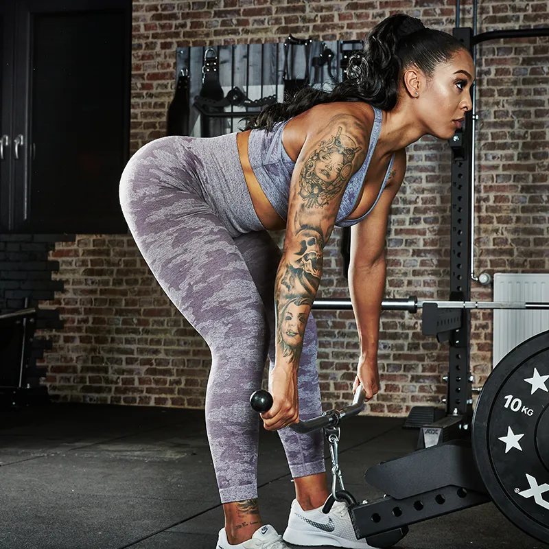 A woman in a gym uses the ATX® Belt Squat Option, wearing a grey sports bra and leggings. She has arm tattoos and is surrounded by weight plates, dumbbells, and other gym equipment.