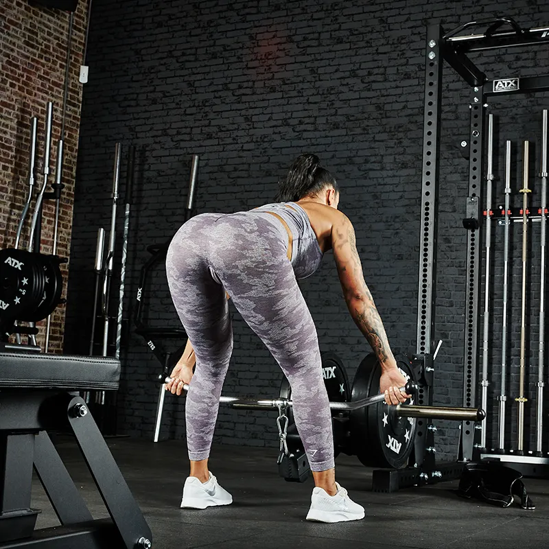 A woman in gray camouflage workout clothes uses the ATX® Belt Squat Option in a gym, standing on a black floor near weight racks and benches, with her back to the camera.