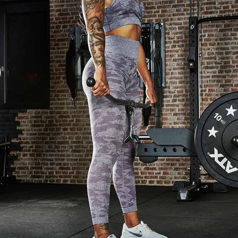 A person in a gym, dressed in a matching purple workout outfit, prepares to exercise with the ATX® Belt Squat Option. Gym equipment and a brick wall can be seen in the background.
