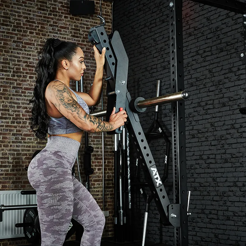 A woman in athletic wear with long dark hair and visible tattoos adjusts the ATX® Belt Squat Option in a gym with brick walls, focused on setting up the equipment.