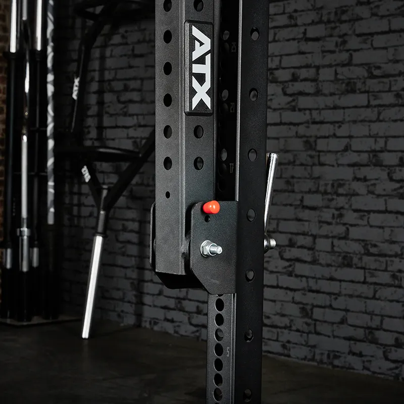 Close-up of an ATX® Belt Squat Option in black, featuring multiple adjustment holes, a safety pin, and a barbell holder, set against a gray brick wall.