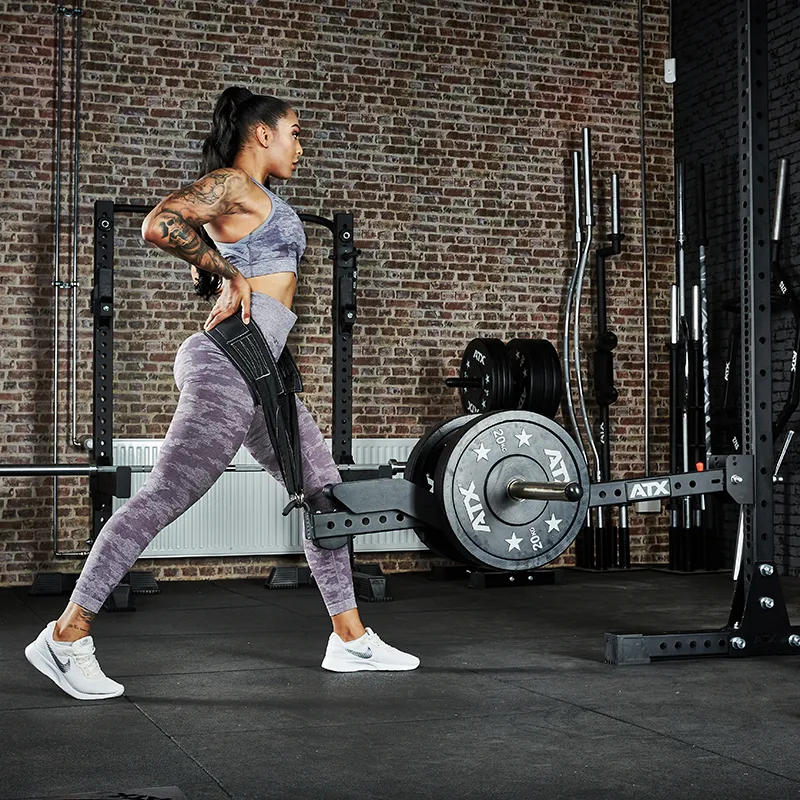 A woman in athletic wear performs a split squat using the ATX® Belt Squat Option on a squat rack in a gym with exposed brick walls and various equipment in the background.