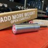 The GymPin 2 Original cylindrical metal weight pin sits on a red surface in a gym, backed by a cardboard box labeled ADD MORE WEIGHT OPEN CAREFULLY, with gym equipment visible behind.