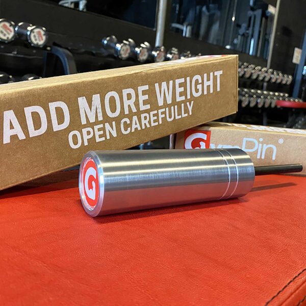 The GymPin 2 Original cylindrical metal weight pin sits on a red surface in a gym, backed by a cardboard box labeled ADD MORE WEIGHT OPEN CAREFULLY, with gym equipment visible behind.
