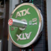 Close-up of a green GymPin 2 Original weight plate mounted on a gym machine, labeled 10 KG, featuring a sleek design with a shiny metal finish. Nearby weights and machine parts are visible in the background.