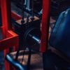 A close-up of a gym machine showcases a sleek black leather seat and red metal frame, with weight plates easily adjusted using the GymPin 2 Original, and numbers visible on the dimly lit weight stack.