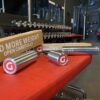 Cylindrical weights with red and white logos rest on a red bench alongside an open box labeled ADD MORE WEIGHT, showcasing a new GymPin 2 Original. In the background, a gleaming rack of silver dumbbells reflects off the mirror.