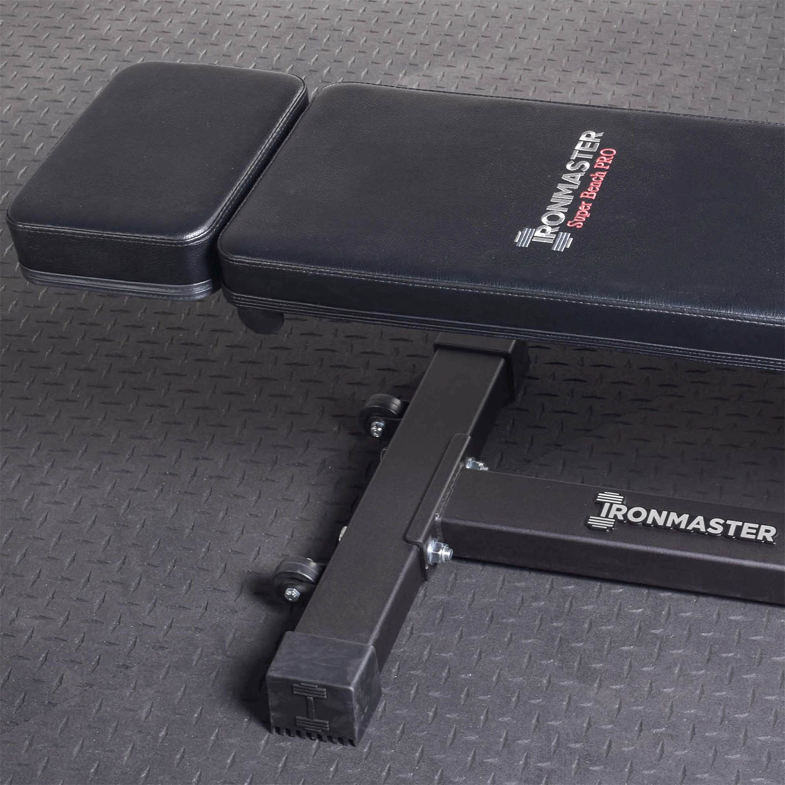 Close-up of a black adjustable gym bench branded Ironmaster Head Extension SB visible on the seat, placed on a textured gray floor.