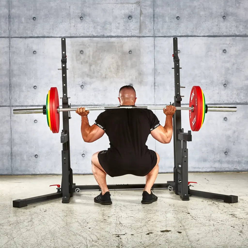A person in black workout attire is executing a squat with a barbell loaded with red and green weight plates on an IPF combo rack. Positioned against a gray industrial-style background, they focus intensely on their form.