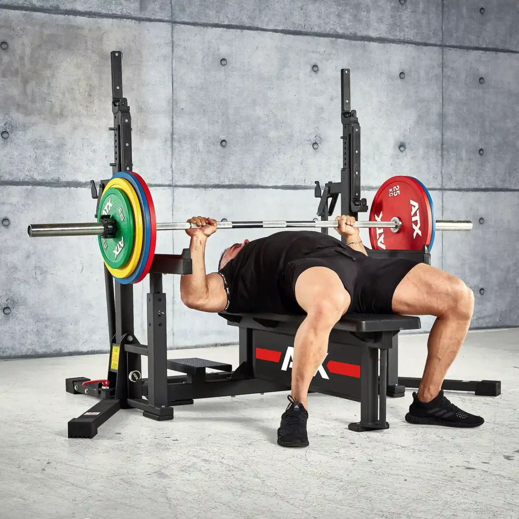 A person is performing a competition bench press on a sturdy squat rack and bench in the gym, lifting a barbell loaded with multicolored weight plates. The background features a concrete wall. The person is wearing a black outfit and sports shoes.