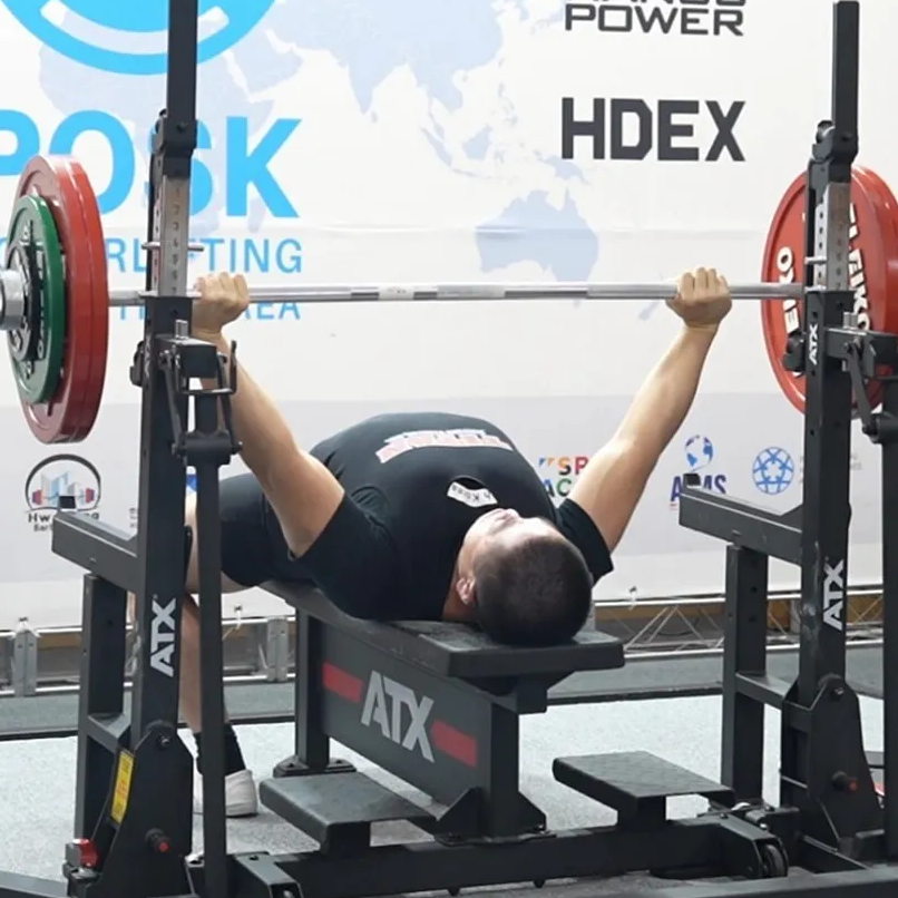 A person is on a bench press gripping a loaded barbell with red and green weights on the ATX® Combo Rack IPF Approved, preparing to lift, with gym equipment and branding visible in the background.