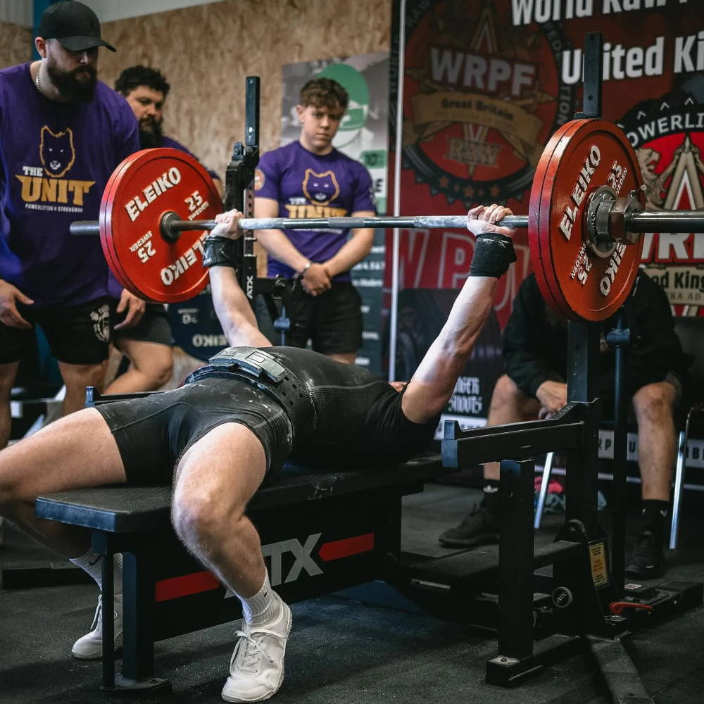 A powerlifter bench presses a loaded barbell on the ATX® Combo Rack IPF Approved during a gym competition, with spotters and onlookers in purple shirts featuring The Unity logo standing nearby, ready to assist.