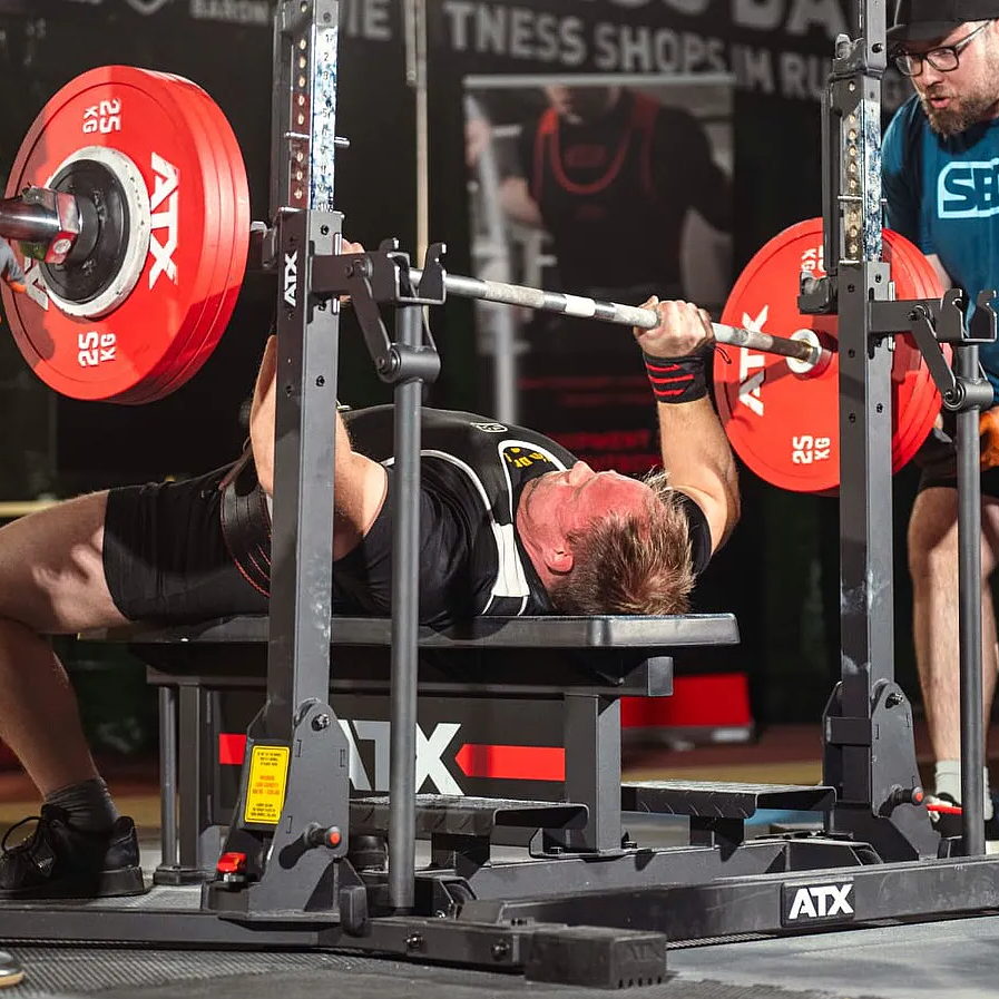 A powerlifter lies on the ATX® Combo Rack IPF Approved, gripping a loaded barbell with red plates, ready to bench press during competition as a spotter stands by for support.