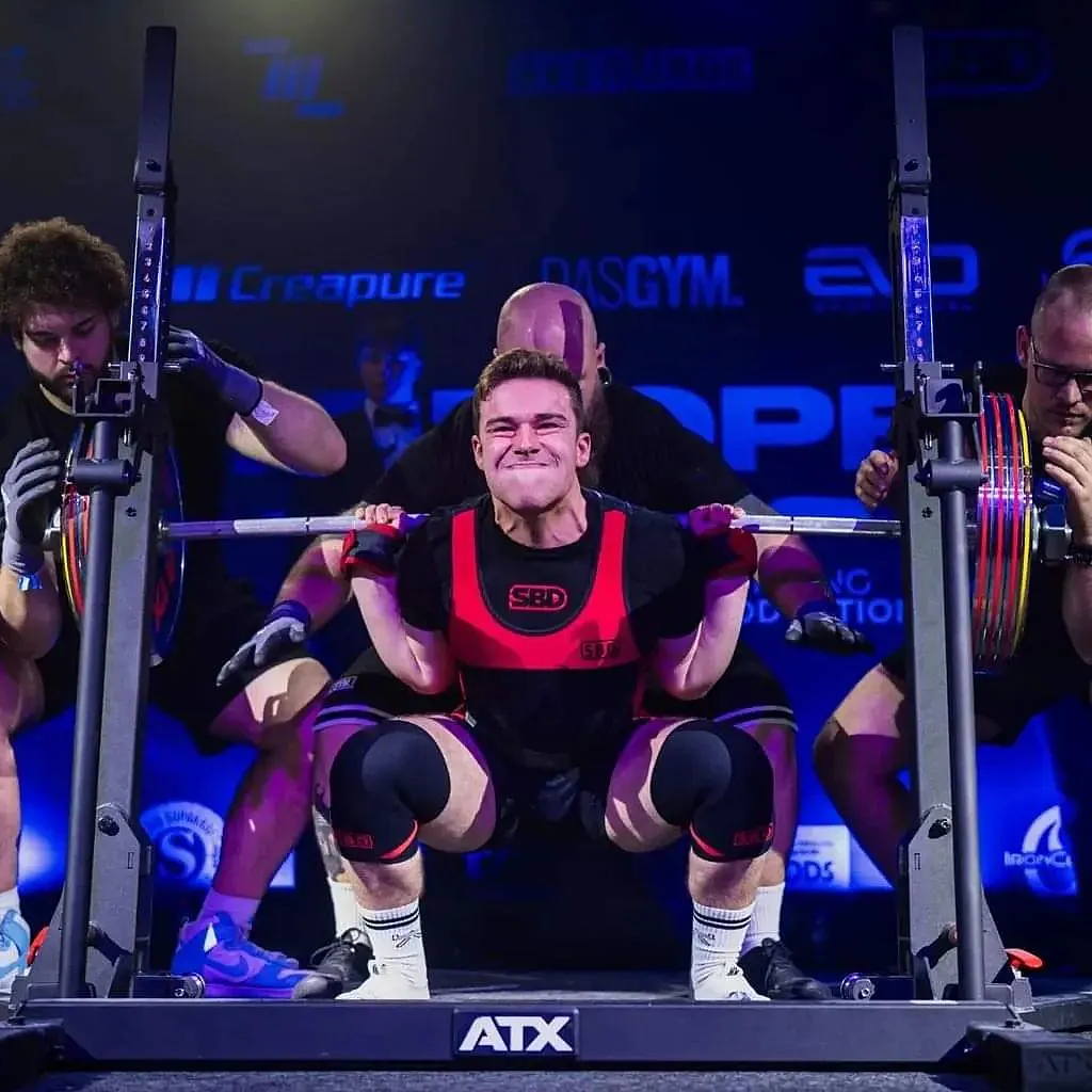 A male powerlifter in a red and black suit strains to perform a heavy squat on the ATX® Combo Rack IPF Approved, surrounded by spotters and loaded weight plates, with event logos and dramatic lighting in the background.