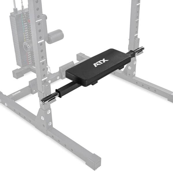 A gym setup includes a black bench pad with ATX printed on it, attached to an adjustable cable crossover machine. The ATX® Hip Thruster Attachment complements the sturdy metal frame and visible pulley system in the background.