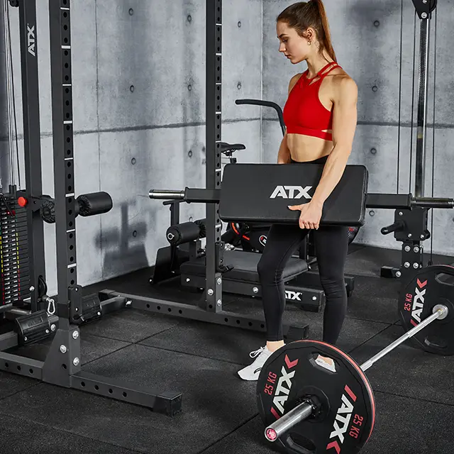A person in a gym holding an ATX® Hip Thruster Attachment rectangular barbell pad in front of a power rack with an attached barbell and weight plates on the floor.