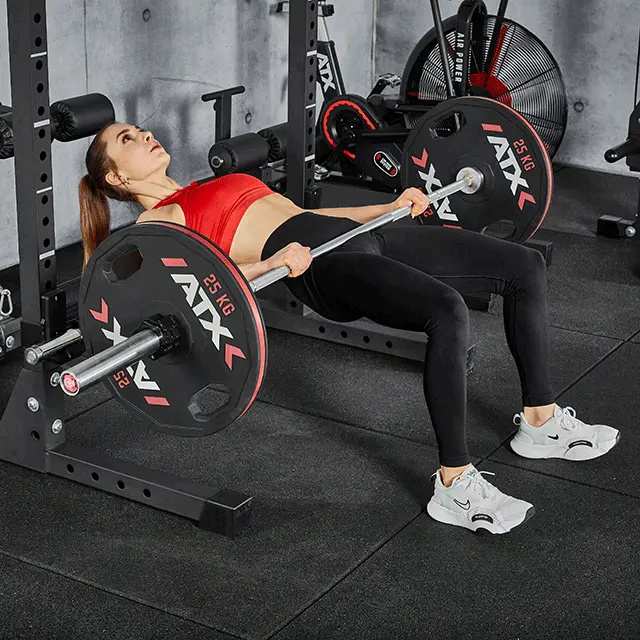A woman performs a hip thrust exercise using an ATX® Hip Thruster Attachment with a barbell loaded with weight plates.