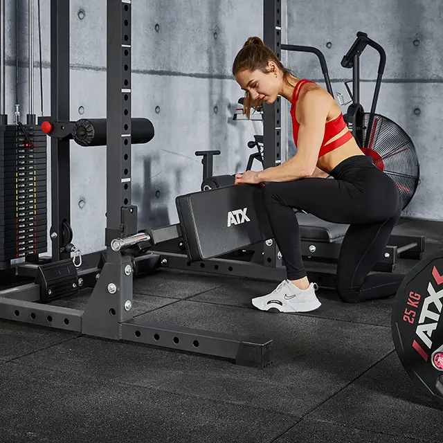 A woman in athletic wear kneels beside a weight rack, adjusting an ATX® Hip Thruster Attachment secured to the frame.