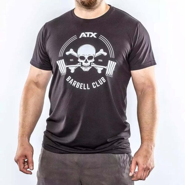 Someone is wearing an ATX® Barbell Club T-Shirt in black, featuring a skull with barbells and the text ATX Barbell Club, along with Est. 1986 above the skull, against a plain white background.