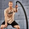 A muscular man exercises with battle ropes in a gym setting. He wears a beige ATX t-shirt and black shorts, with a focused expression. The concrete wall background adds to the rugged, intense workout atmosphere.