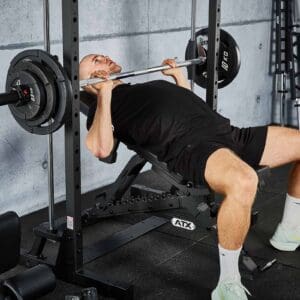 In a gym with concrete walls, a person in black is using the ATX® Smith Machine 500 (198 cm) for bench pressing, surrounded by fitness equipment.