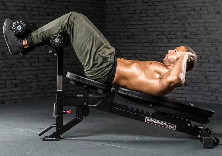 A man in athletic gear performs decline sit-ups on a workout bench with the ATX® AB Attachment, gripping his head with both hands and securing his feet under padded rollers in a gym with dark brick walls.