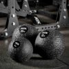Three ATX® Slam Balls in black texture (4 kg, 12 kg, 20 kg) rest on the gym floor with a metal exercise rack seen behind.